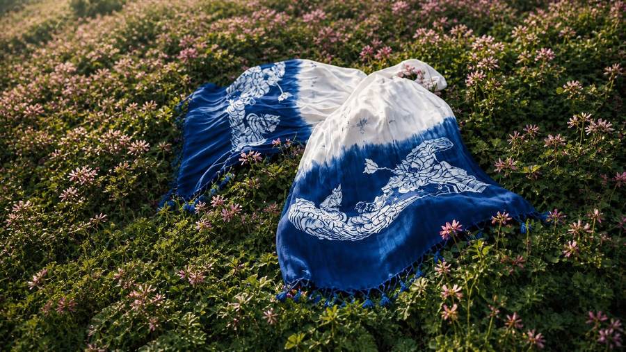 An indigo-dyed scarf with a white pattern and knotted tassels lying on a hillside of pink flowers