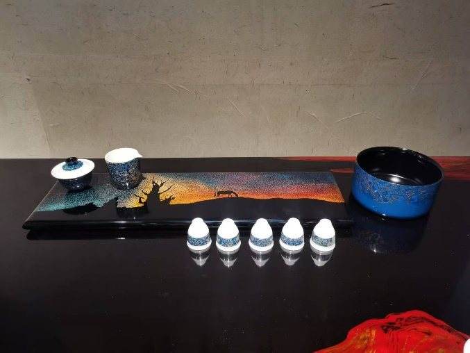 A black lacquer tea tray with a sunset landscape, small cups and a blue bowl