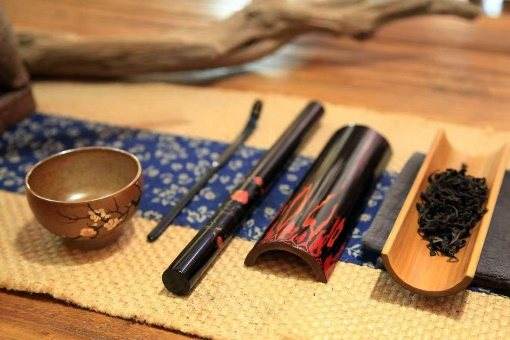 Black lacquer tea tools with red flame patterns beside a wooden cup and a bamboo scoop of tea leaves