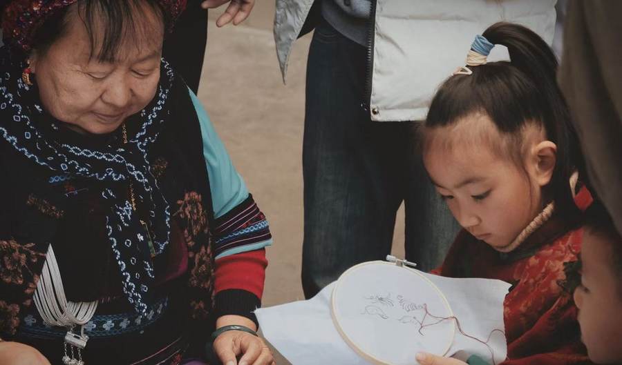 An elder woman in embroidered dress and silver necklace guides a child holding an embroidery hoop