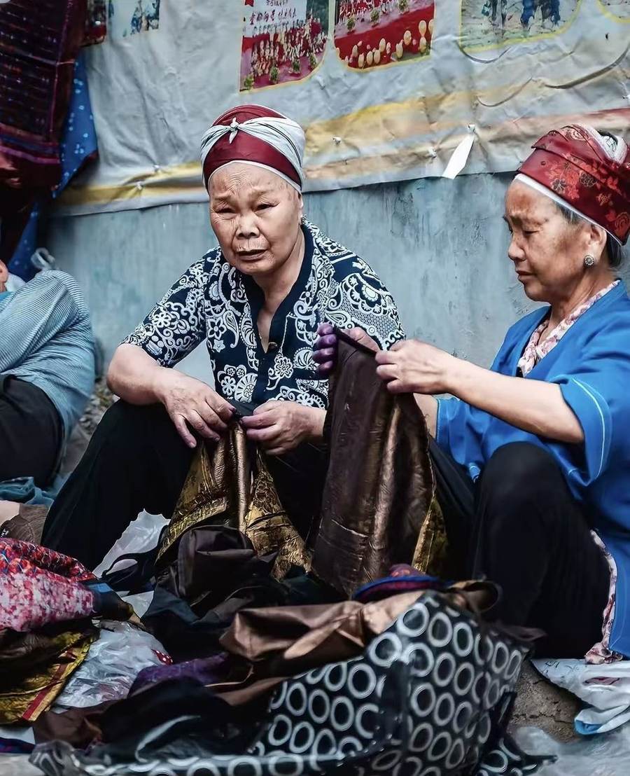 Two elder women in headwraps sorting bundles of patterned and shimmering cloth at a market