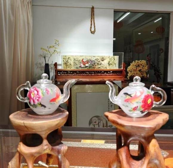 Two small clear glass teapots on wooden stands, painted with a pink flower and an orange fish