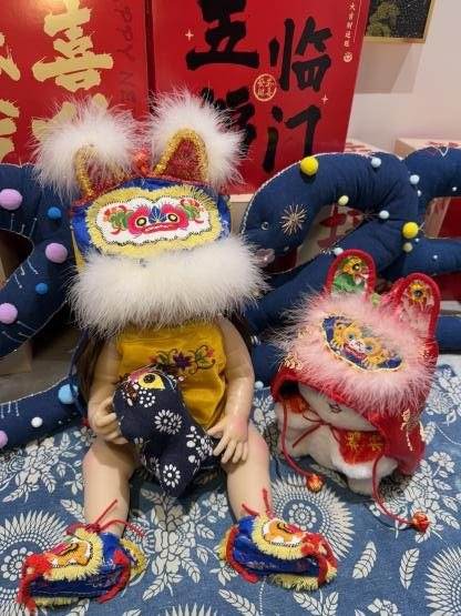 A doll in a tiger hat and tiger shoes, beside a second doll in a red tiger hat, on blue printed cloth
